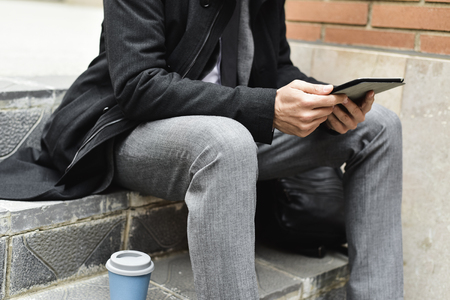 Closeup of a young caucasian businessman in a gray suit and a gray coat sitting on a outdoors staircase using a tablet next to a cup of coffeeの写真素材