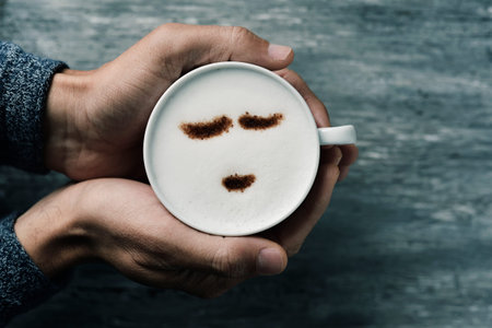 high-angle shot of a young caucasian man holding a cup of cappuccino with a happy face drawn with cocoa powder on the milk foam, on a gray rustic tableの写真素材