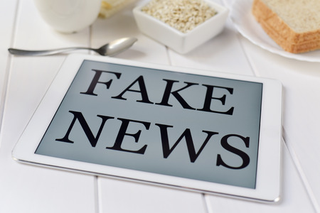 closeup of a tablet computer with the text fake news in its screen on a white table set for breakfast, with some cereal, a cup of coffee and some slices of breadの写真素材