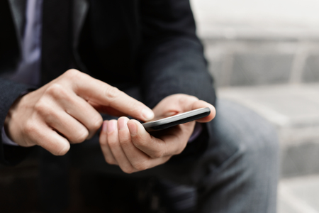 closeup of a young caucasian businessman in a gray suit and a dark gray coat using a smartphone outdoorsの写真素材