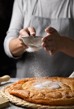 closeup of a young caucasian man sprinkling powdered sugar on an ensaimada, a pastry typical of Mallorca, Spain, placed on a rustic wooden tableの写真素材