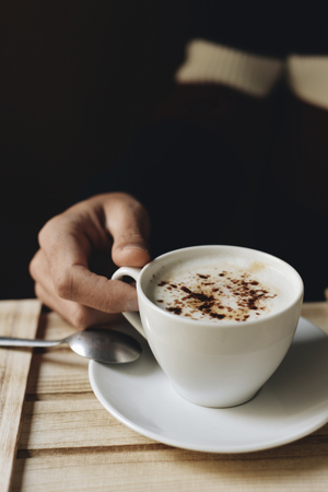 closeup of a young caucasian man about to drink a cup of cappuccino, sitting at a rustic wooden tableの写真素材