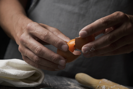 closeup of a young caucasian man separating the yolk of an egg using the broken shell, on a rustic wooden table next to a rolling pinの写真素材
