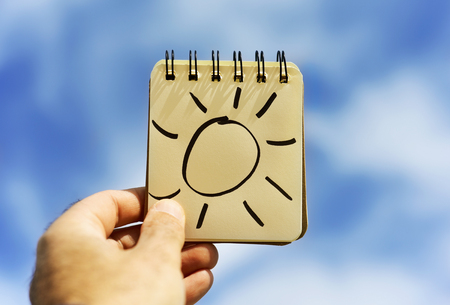 closeup of a young caucasian man holding a spiral notepad with a sun drawn in it against the skyの写真素材