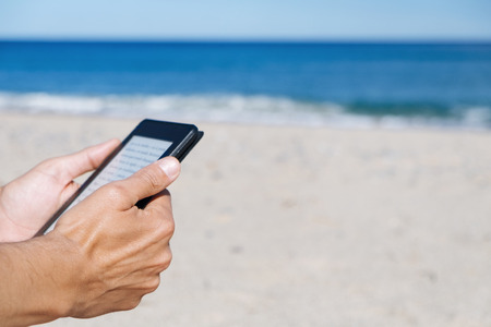 closeup of a young caucasian man reading in a tablet or e-reader on a white sand beach next to the seaの写真素材