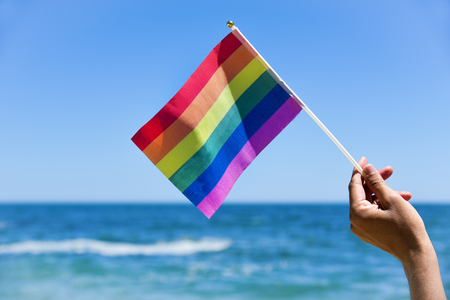 closeup of a young caucasian man waving a small rainbow flag in front of the oceanの写真素材