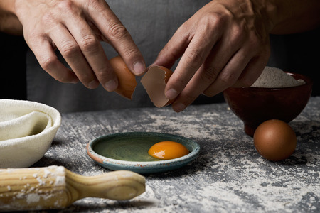 closeup of a young caucasian man wearing a gray apron cracking an egg on a rustic wooden table sprinkled with flour next to a rolling pinの写真素材