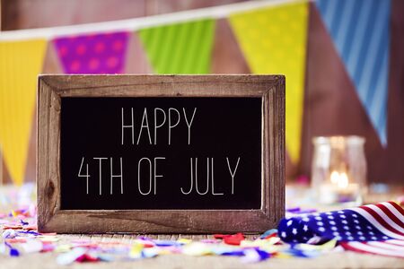 a wooden-framed chalkboard with the text happy 4th of july written in it and an American flag, placed on a rustic wooden background sprinkled with confetti and a colorful garland in the backgroundの写真素材