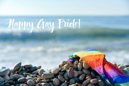 closeup of a rolled rainbow flag in a shingle beach, with the ocean in the background, and the text happy gay prideの写真素材