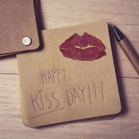high-angle shot of a brown note with the text happy kiss day written in it and the mark of a pair of red lips, on a wooden table next to a penの写真素材