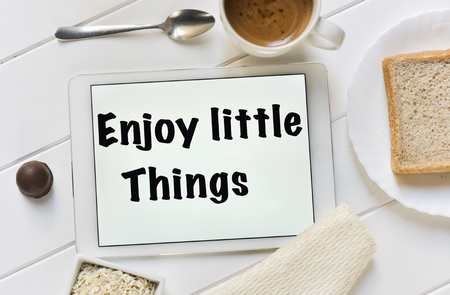 high-angle shot of a tablet with the text enjoy little things in its screen and a cup of coffee, a bowl with cereals and a plate with a slice of bread on a white table set for breakfastの写真素材