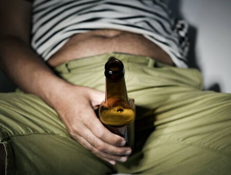 closeup of a young caucasian man with a bottle of beer in his hand sitting in the sofa, showing his hairy bellyの写真素材