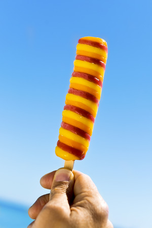 closeup of a refreshing popsicle in the hand of a young caucasian man against the blue sky, with the sea in the backgroundの写真素材