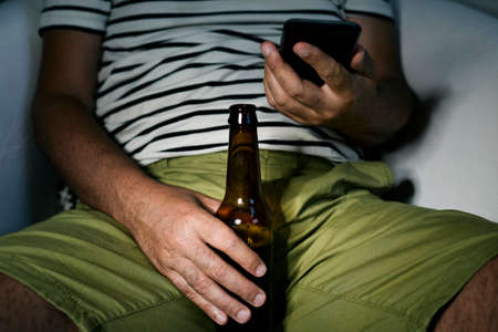 closeup of a young caucasian man drinking a beer and using his smartphone, sitting in a sofaの写真素材