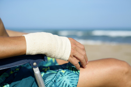 closeup of a young caucasian man in swimsuit sitting in a deck chair on the beach with a bandage in his wristの写真素材