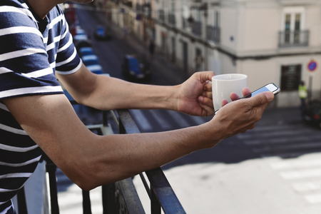 closeup of a young caucasian man drinking coffee and using his smartphone in the balcony of his home or of a hotelの写真素材
