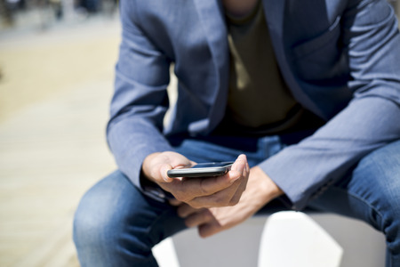 closeup of a young caucasian man, wearing a smart casual look, a pair of jeans and an elegant blue jacket, using a smartphone next to the beachの写真素材