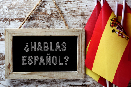 a wooden-framed chalkboard with the question hablas espanol? do you speak Spanish? written in Spanish, and some flags of Spain against a rustic wooden backgroundの写真素材