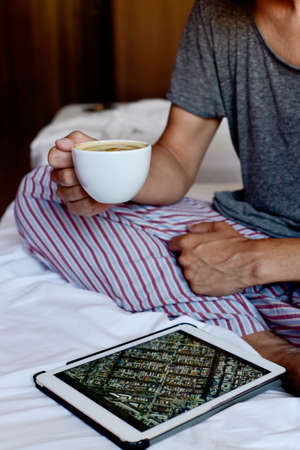 closeup of a young caucasian man in pajamas sitting in bed having a coffee while is checking an address in a map in his tabletの写真素材