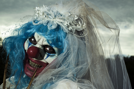 closeup of a scary evil clown wearing a dirty and ragged bride dress, in a disturbing landscape at nightの写真素材