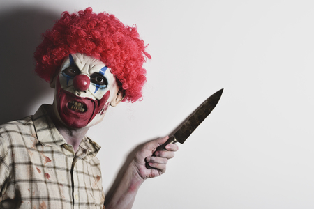 a scary evil clown wearing a dirty and ragged plaid shirt with a big knife in his hand against an off-white background with a blank space on the rightの写真素材