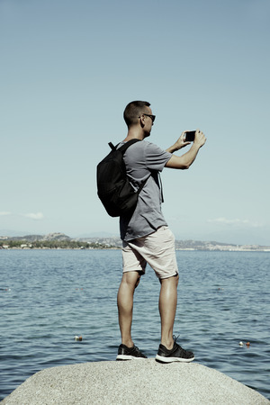 closeup of a young caucasian man carrying a backpack taking a photo of the sea in the Costa Smeralda, in Sardinia, Italy, with his smartphoneの写真素材