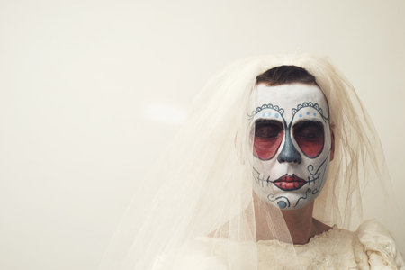 closeup of a person with short hair in a bride dress with a mexican calaveras makeup, wearing veil, against an off-white background with a blank space on the leftの写真素材