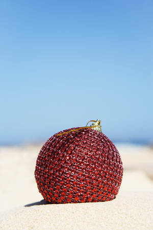 closeup of an elegant red christmas ball in the sand of a beach with the sky in the background and a large blank space on topの写真素材