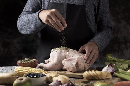 closeup of a young caucasian man preparing a turkey placed on a rustic wooden table full of ingredients to stuff it such as apple, onion or different spices, and celery or a roast corn on the cobの写真素材