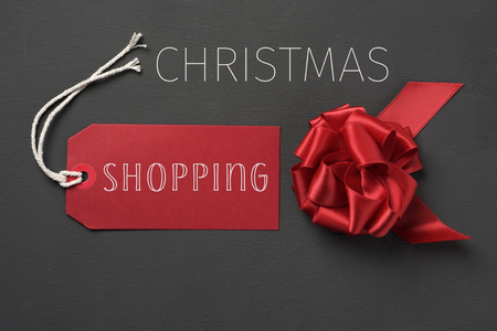 the words christmas and shopping in a red paper label and a red satin ribbon bow, against a dark gray backgroundの写真素材