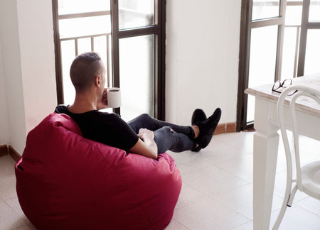 a young caucasian man indoors have a coffee sitting in a comfortable red bean bag chairの写真素材
