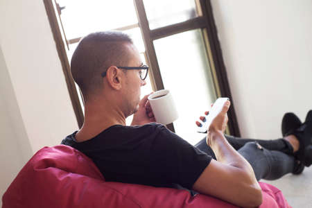 a young caucasian man indoors have a coffee while is using his smartphone sitting in a comfortable red bean bag chairの写真素材