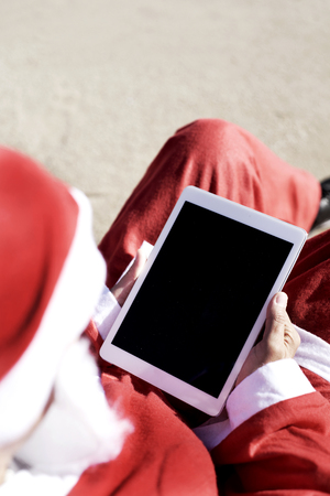 closeup of santa claus using his tablet, with a black blank space in its screen, sitting on the beach の写真素材