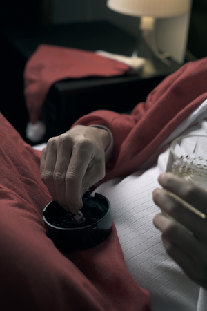 closeup of a young man wearing a santa suit drinking whisky and smoking a cigarette in bedの写真素材
