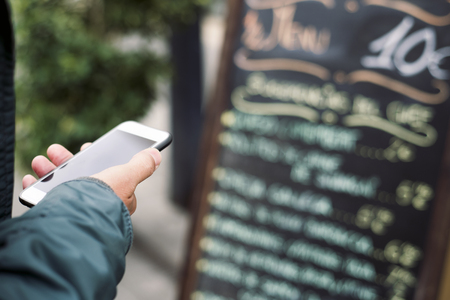 closeup of a young caucasian man using a smartphone in the street, in front of a restaurant, reading the online reviews of the premises or reserving a tableの写真素材