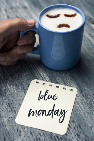 closeup of a note with the text blue Monday written in it on a gray rustic table and a young caucasian man with a blue cup of cappuccino with a sad face drawn with cocoa powder on its milk foamの写真素材