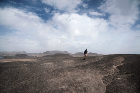 a young caucasian man, seen from behind, carrying a backpack walking by a volcanic landscape in...