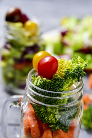 a salad with a mix of different vegetables, such as cauliflower, carrot, broccoli and cherry tomatoes of different colors served in a mason jar, on a gray rustic wooden tableの写真素材