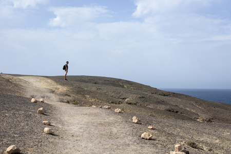 a young caucasian man, seen from behind, carrying a backpack walking by a volcanic landscape in Fuerteventura, Canary Islands, Spainの写真素材