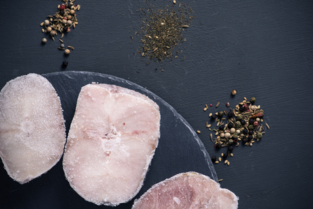high angle view of some frozen hake cuts on a round slate tray, placed on a dark gray surface with some piles of different spices, such as different kinds of peppercornsの写真素材