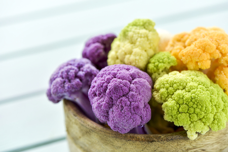 closeup of a rustic wooden bowl with some pieces of cauliflower of different colors, such as green, purple and orange, on a pale green tableの写真素材