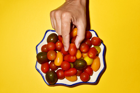 High angle view of the hand of a young caucasian man picking a tomato from a plate, in which there is an assortment of different cherry tomatoes, placed on a bright yellow backgroundの写真素材