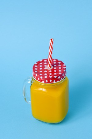an orange beverage served in a glass mason jar with a red lid patterned with white dots, and a red and white drawing straw, on a blue background with some blank space on topの写真素材
