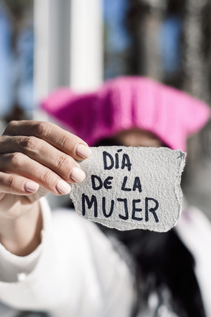 closeup of a young caucasian woman outdoors wearing a pink hat showing a piece of paper in front of her face with the text dia de la mujer, womens day written in spanishの写真素材