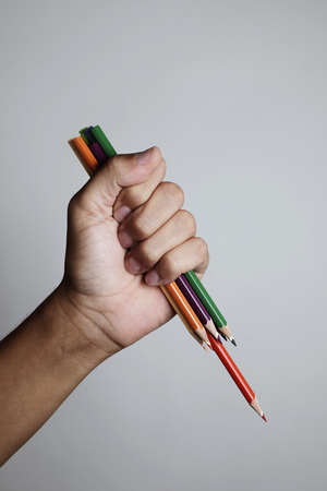 closeup of a young caucasian man with a bunch of sharpened pencil crayons in his hand in a threatening gesture like wielding a knife against an off-white background, with some blank space on topの写真素材