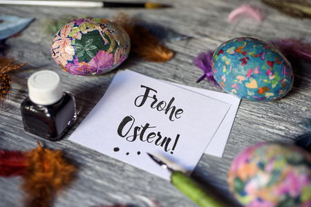 closeup of a piece of paper with the text frohe ostern, happy easter written in german, an ink bottle, a dip pen, some different homemade easter eggs and some feathers, on a gray rustic wooden tableの写真素材