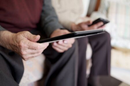 closeup of an old caucasian man using a tablet and an old caucasian man using a smartphone, sitting both in a couchの写真素材