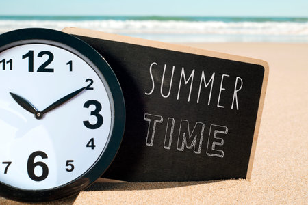 closeup of a clock and a signboard with the text summer time written in it, on the sand of a beach
