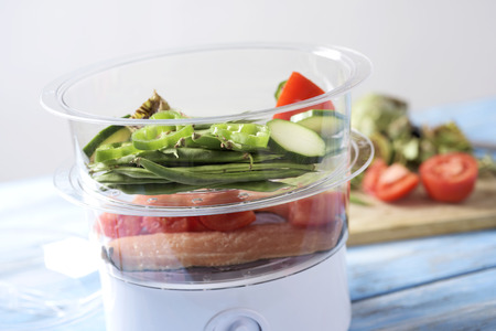 closeup of an electric steam cooker with some raw food, such as green beans, zucchini, green pepper, tomato or fish, on a pale blue wooden tableの写真素材