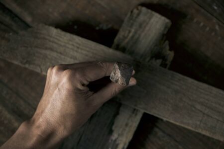 closeup of a young man driving a rusty nail on a wooden crossの写真素材